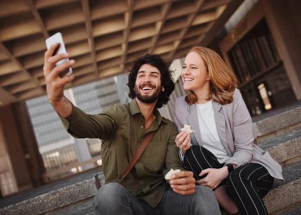 Mutlu profesyonel çift, öğle arasında sandviçlerini yerken akıllı telefon kullanarak selfie çekiyor. — Stok fotoğraf