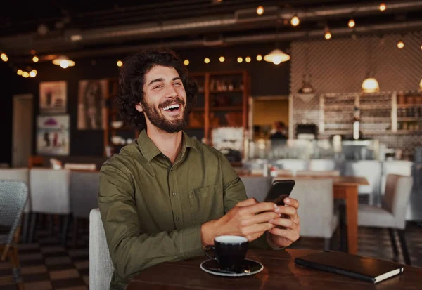 Porträt eines gut aussehenden jungen Mannes, der lacht, während er in der Freizeit in der Cafeteria sitzt und eine lustige Nachricht am Telefon liest — Stockfoto