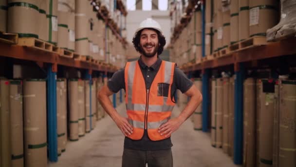 Jovem trabalhador sorridente de pé com os braços na cintura em um grande armazém com pilha de produtos manufaturados — Vídeo de Stock