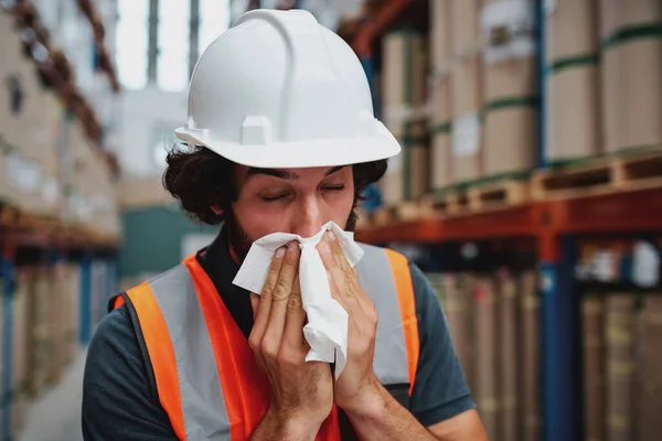 Giovane magazziniere si sente male mentre starnutisce e tossisce in fabbrica in piedi vicino a scatole di cartone imballate sullo scaffale — Foto Stock