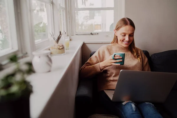 Glimlachende jonge vrouw onder zelfisolatie genieten van 's morgens koffie met behulp van laptop zitten thuis op de bank — Stockfoto
