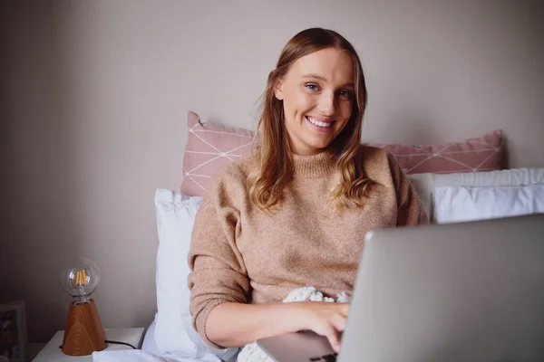 Freelancer feminino feliz sentar na cama moderna e digitando laptop durante o período de bloqueio do vírus corona sorrindo para a câmera — Fotografia de Stock