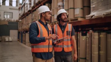 İki çok ırklı erkek işçi büyük bir depoda dikilip dijital tablet üzerinde bir kontrol listesi hazırlıyorlar.