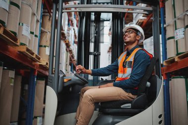 Afrikalı forklift sürücüsü beyaz kasklı ve yelekli büyük bir depodaki stokları dikkatlice taşımaya odaklanmıştı.