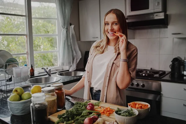 Cheerful young woman in modern kitchen preparing fresh vegetables salad and eating a carrot smiling at the camera — Stock Photo, Image