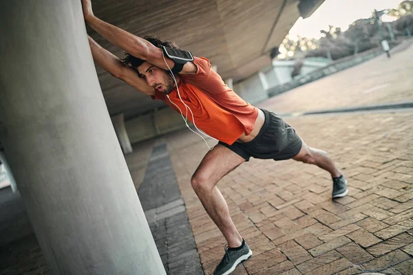 Retrato de un hombre de fitness con brazalete y auricular haciendo ejercicios de estiramiento bajo el puente —  Fotos de Stock