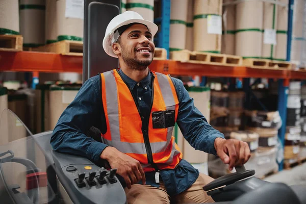 Happy forklift sürücüsü beyaz kask ve yelek giyerek büyük bir deponun zeminindeki raflardan stokları dikkatlice taşımaya odaklanmış.