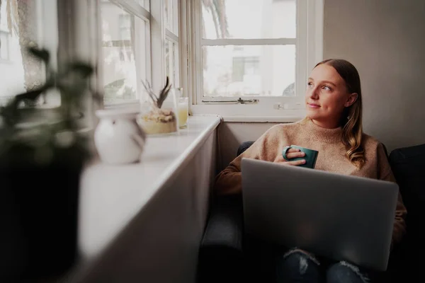 自宅でコーヒーカップを手に窓の外を見て座っているノートパソコンで働く思慮深い若いビジネスマンの女性 — ストック写真