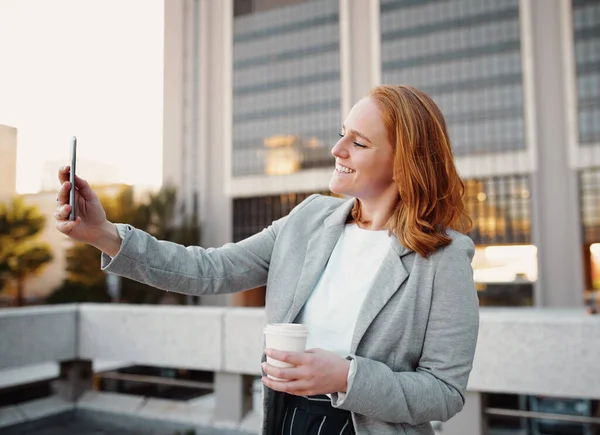 Mulher de negócios sorridente em roupas formais segurando taça de café takeaway de pé ao ar livre enquanto toma selfie usando smartphone — Fotografia de Stock