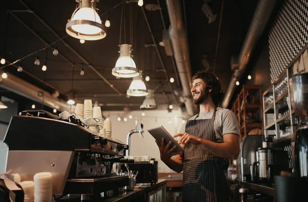 Smiling caucasian man wearing apron standing behind cafe counter using digital tablet looking away — Stock Photo, Image