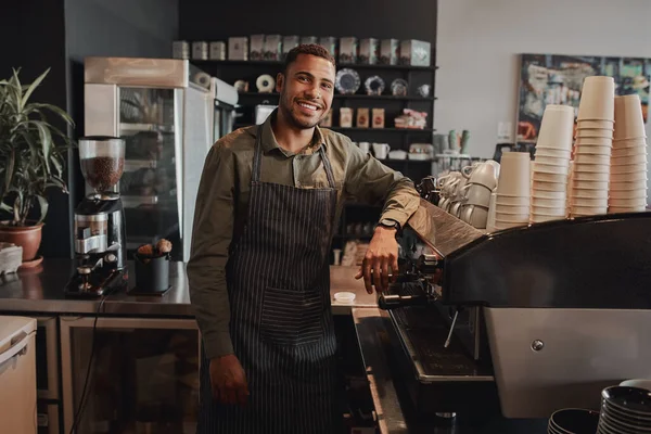 Succesvolle afro-Amerikaanse mannelijke ondernemer achter de toonbank van een coffeeshop leunend aan de toonbank glimlachend kijkend naar de camera — Stockfoto