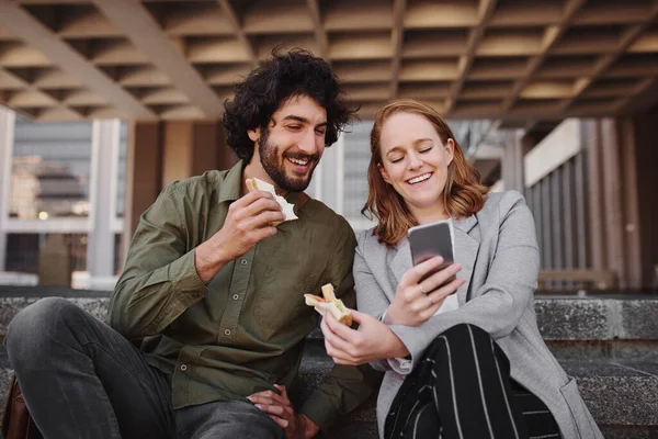 Casal assistindo vídeo no smartphone enquanto come sanduíche durante a pausa do escritório — Fotografia de Stock