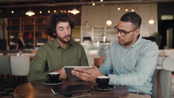 Hombre de negocios multiétnico que muestra la presentación al cliente en la cafetería usando tableta digital mientras bebe café: dos hombres mirando una tableta — Vídeos de Stock