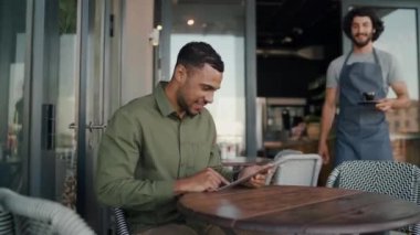 Afrikalı müşteriye kahve servisi yapan genç, mutlu, beyaz bir garson dışarıda oturmuş dijital tablet kullanıyor.
