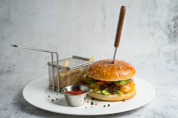 Big tasty burger with knife in it served with tomato souse and french fries on a grey background