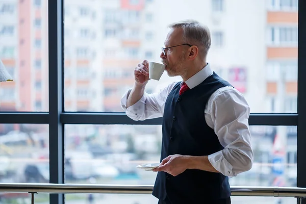 Smiling happy managing director thinks about his successful career development while standing with a cup of coffee in his hand in his office near the background of a window with copy space
