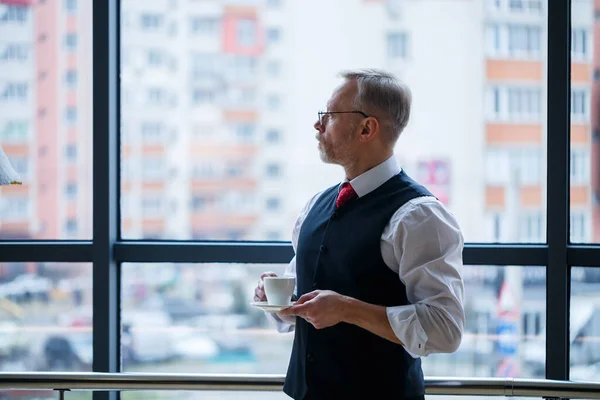 Smiling happy managing director thinks about his successful career development while standing with a cup of coffee in his hand in his office near the background of a window with copy space