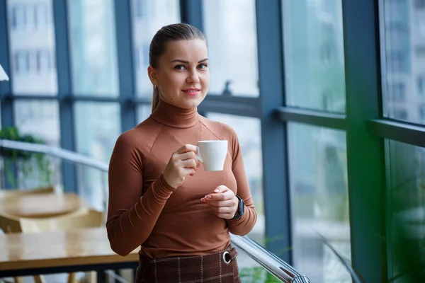 Smiling happy managing woman director thinking about her successful career development while standing with a cup of aromatic coffee in her office near the background of a window with copy space