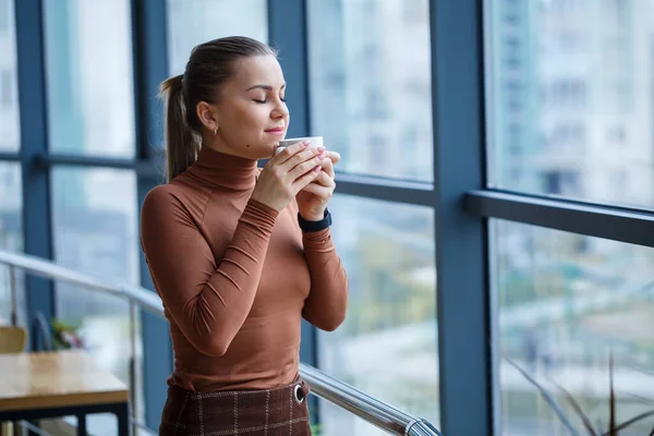 Smiling happy managing woman director thinking about her successful career development while standing with a cup of aromatic coffee in her office near the background of a window with copy space