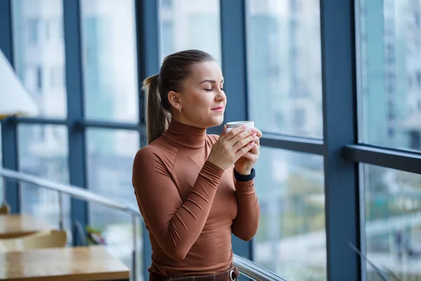 Smiling happy managing woman director thinking about her successful career development while standing with a cup of aromatic coffee in her office near the background of a window with copy space