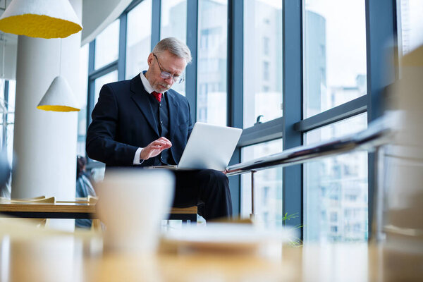 Smiling happy managing director thinks about his successful career development while standing with a laptop in his office near the background of a window with copy space