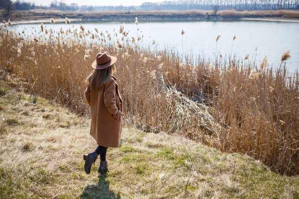 茶色のコートを着た女の子 帽子と眼鏡は明るい太陽の下で湖と公園を歩いています 人生と笑顔の喜び 春の始まり — ストック写真