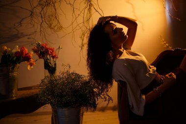 Kahverengi ışıklı bir kız kanepede oturuyor. Vazodaki taze çiçeklerin etrafında.