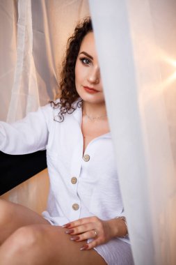 Hassas, bronz tenli, neşeli, narin, dalgalı saçlı, sandalyede oturan, beyaz tül, açık renk kumaş, beyaz gömlek, çıplak bacaklar, seksi, çekici.