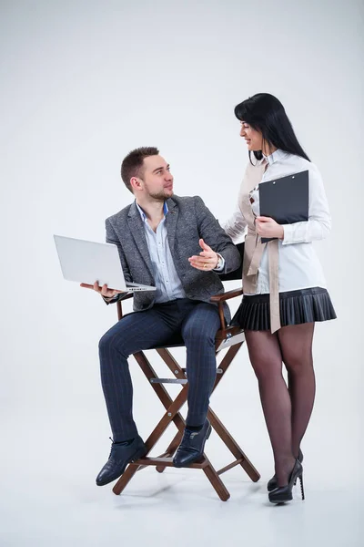 A successful man in a suit is sitting on a black chair with a laptop and a woman is standing nearby. Future businessmen are smiling and talking about a new project.