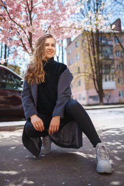 Uzun dalgalı saçlı, gri paltolu, spor ayakkabılı genç bir kadın model sokakta oturuyor. Bahar ağaçları kadın gülüyor ve seviniyor.