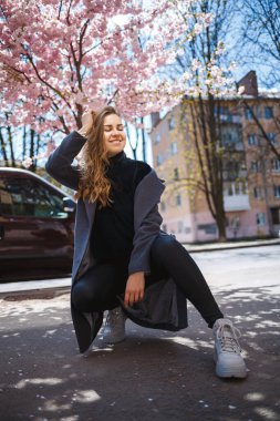 Uzun dalgalı saçlı, gri paltolu, spor ayakkabılı genç bir kadın model sokakta oturuyor. Bahar ağaçları kadın gülüyor ve seviniyor.