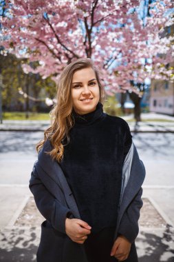 Uzun dalgalı saçlı, gri paltolu, spor ayakkabılı genç bir kadın model, arka planda güzel pembe çiçeklerle çiçekli bir çalılığın yanında duruyor ve poz veriyor..