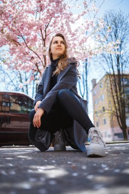 Uzun dalgalı saçlı, gri paltolu, spor ayakkabılı genç bir kadın model sokakta oturuyor. Bahar ağaçları kadın gülüyor ve seviniyor.