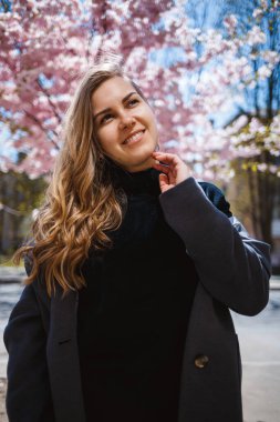 Uzun dalgalı saçlı, gri paltolu, spor ayakkabılı genç bir kadın model, arka planda güzel pembe çiçeklerle çiçekli bir çalılığın yanında duruyor ve poz veriyor..