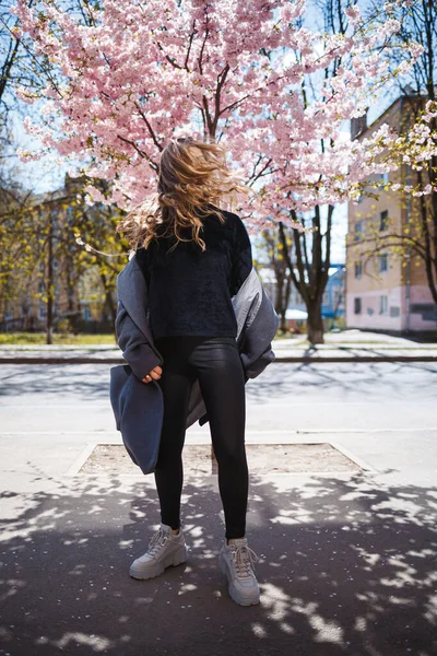 長い波状の髪を持つ若いスレンダーな女性モデルと 灰色のコート スニーカーで身を包んだ 通りに回転 春の花の木の女の子は笑うと喜びを実行します — ストック写真