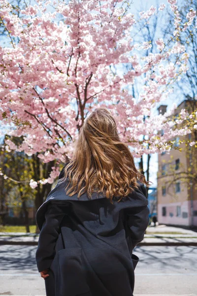 長い波状の髪を持つ若いスレンダーな女性モデルと 灰色のコート スニーカーで身を包んだ 通りに回転 春の花の木の女の子は笑うと喜びを実行します — ストック写真