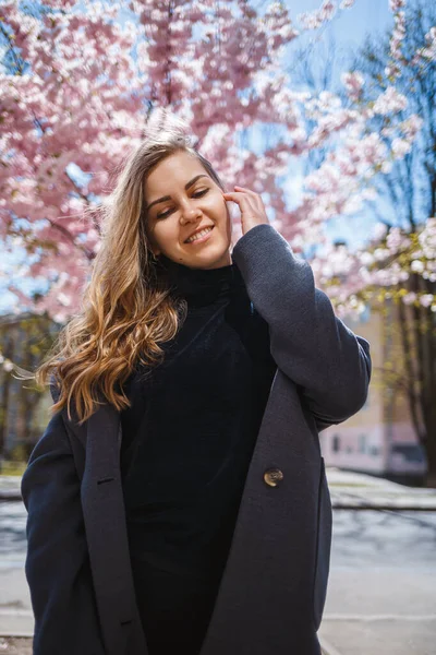 長い波状の髪をした若いスレンダーな女性モデルで 灰色のコート スニーカーを身に着けて 背景に美しいピンクの花を持つ開花低木の近くの通りに立ち ポーズ — ストック写真