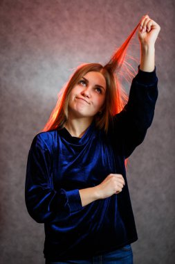 Sarı saçlı, duygusal olarak gri arka planda poz veren bir kız. Saçları kırmızıyla aydınlatılmış.