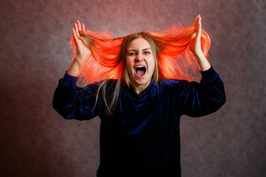 Sarı saçlı, duygusal olarak gri arka planda poz veren bir kız. Saçları kırmızıyla aydınlatılmış.