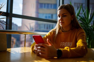 Sarı kazaklı kız büyük bir pencerenin yanında oturuyor ve kırmızı bir telefonu var.