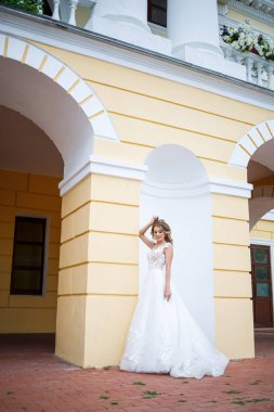 Gelinin düğün gününde, büyük bir evin arka planında beyaz bir gelinlik giymiş, güzel bir genç kızın portresi.