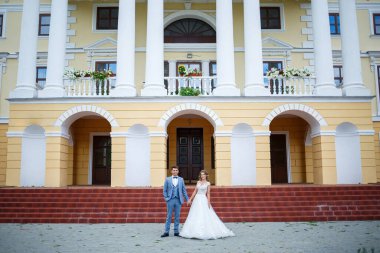 Damat ve gelin kostümü giymiş genç şık bir adam. Beyaz elbiseli güzel bir kız. Düğün günlerinde büyük bir evin arka planında yürüyorlar.