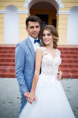 Damat ve gelin kostümü giymiş genç şık bir adam. Beyaz elbiseli güzel bir kız. Düğün günlerinde büyük bir evin arka planında yürüyorlar.