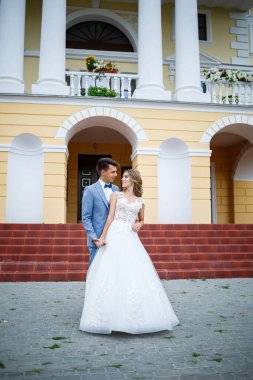 Damat ve gelin kostümü giymiş genç şık bir adam. Beyaz elbiseli güzel bir kız. Düğün günlerinde büyük bir evin arka planında yürüyorlar.