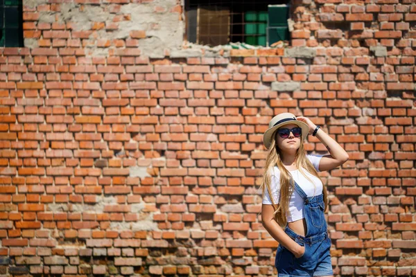 Girl in denim overalls, a white T-shirt and a light hat on a background of a brick building under the bright sun
