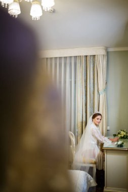 Gelin beyaz önlüklü ve peçeli makyaj masasında oturuyor.