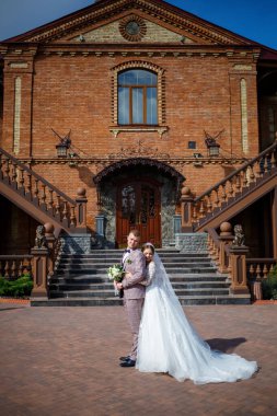 Beyaz gelinlik içinde gelin ve büyük adımlı tuğla bir binanın arka planında damat.