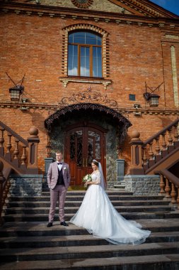 Beyaz gelinlik içinde gelin ve büyük adımlı tuğla bir binanın arka planında damat.