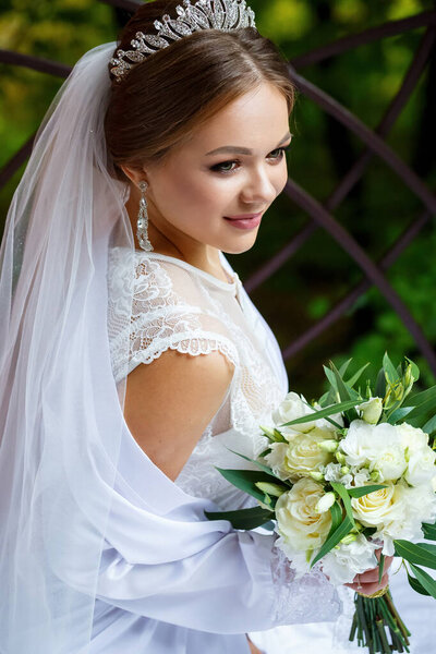 beautiful bride in veil and white coat sits on a blanket with a wedding bouquet in her hands