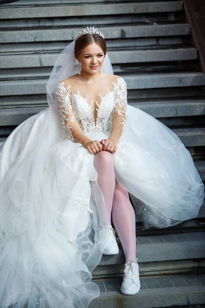 Beyaz gelinlik ve beyaz spor ayakkabılar içinde gelin büyük bir binanın merdivenlerinde oturuyor.